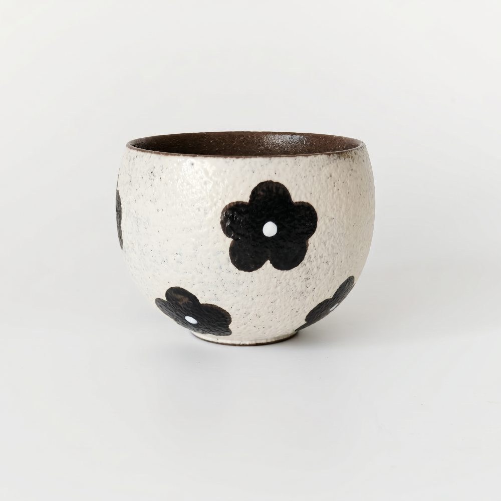 A slightly elevated view of a textured ceramic handleless tea cup by Kove Ceramics, highlighting the striking contrast between its deep dark glazed interior and the speckled cream exterior with bold black floral patterns.