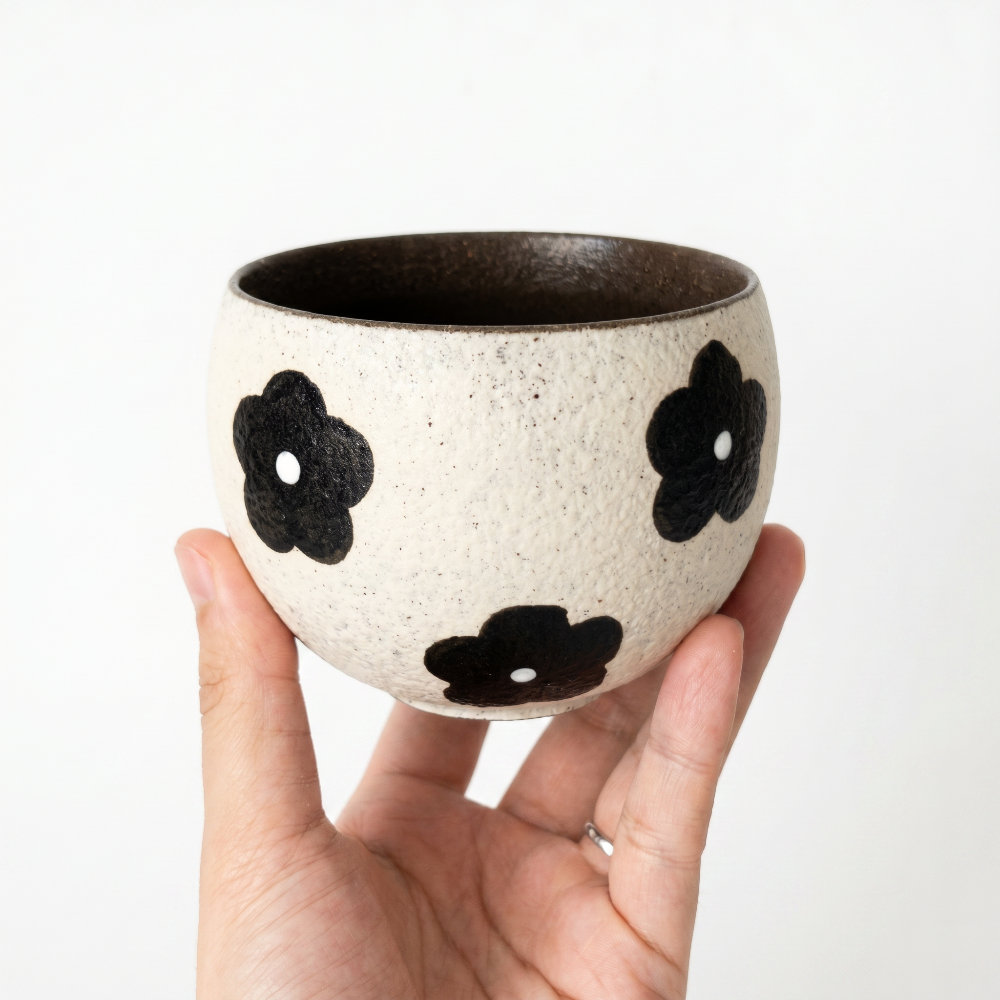 A hand holding a round, textured speckled cream ceramic handleless cup by Kove Ceramics, featuring bold black retro floral patterns and a dark interior, demonstrating its perfect size for matcha or tea.
