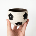 A hand holding a round, textured speckled cream ceramic handleless cup by Kove Ceramics, featuring bold black retro floral patterns and a dark interior, demonstrating its perfect size for matcha or tea.