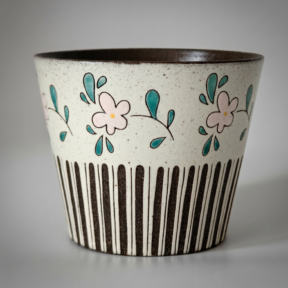 A detailed close-up of a tapered, handmade speckled stoneware cup by Kove Ceramics against a soft grey background. The lighting beautifully highlights the tactile texture of its black vertical stripes, the dark interior glaze, and the delicate hand-painted botanical pink flowers.