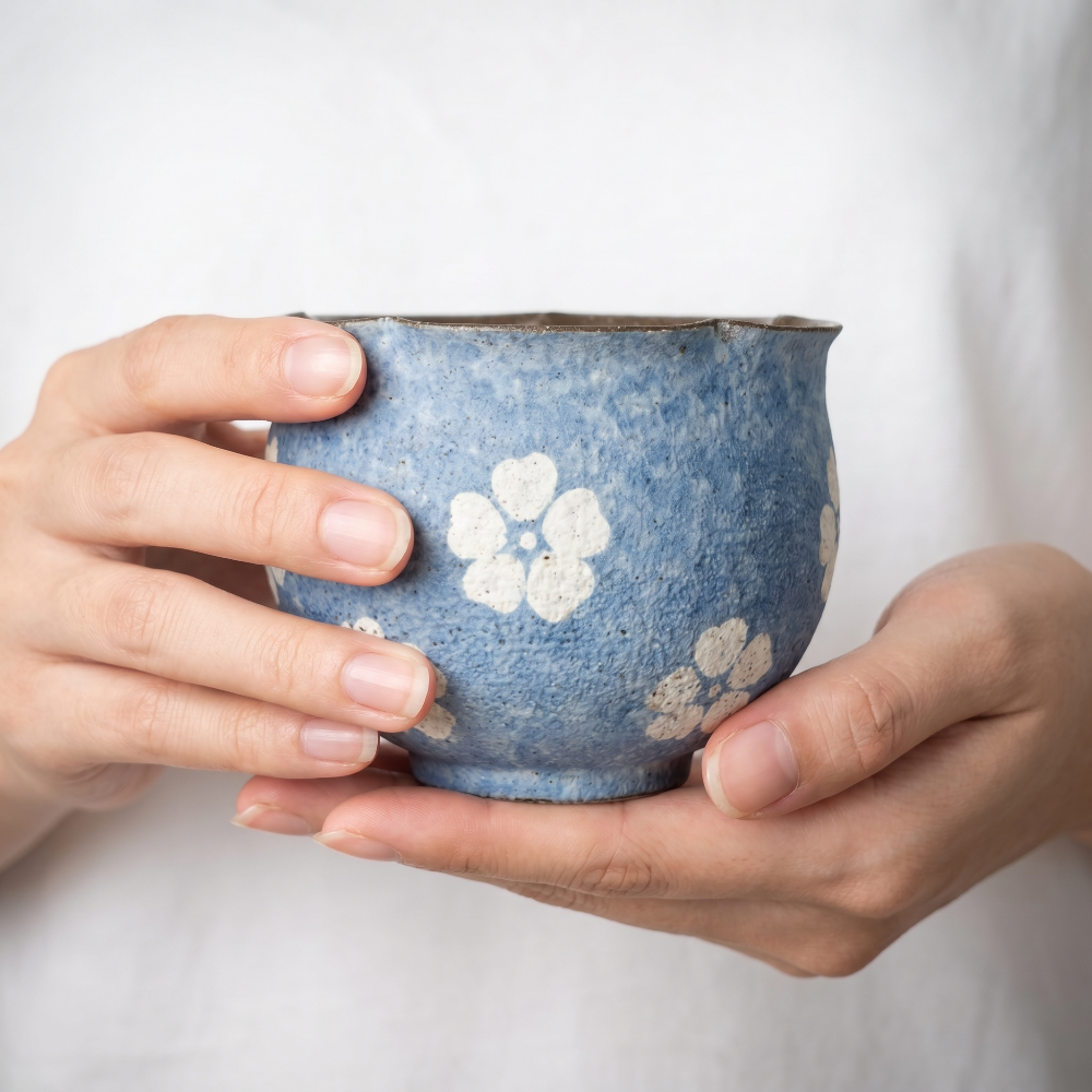 A cozy lifestyle close-up showing two hands gently cupping a handleless ceramic tea bowl by Kove Ceramics against a soft white fabric background. The cup features a textured, mottled blue glaze and hand-painted white blossoms, conveying a warm and comforting aesthetic