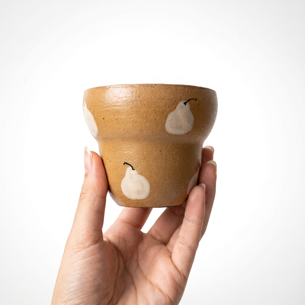 A close-up shot of a hand delicately supporting a stepped ceramic cup by Kove Ceramics from the base. The image highlights the cup's glossy mustard yellow speckled glaze and the hand-painted white pear designs against a bright white background.