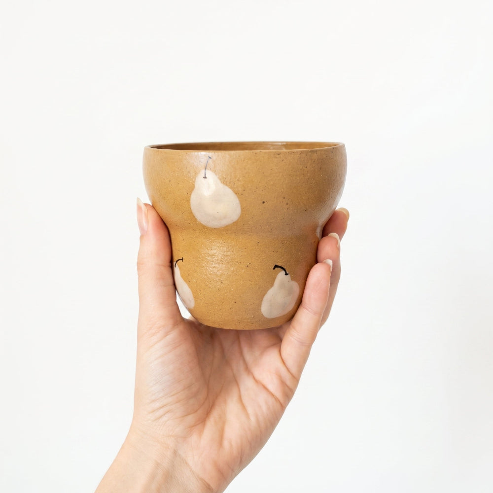A front-view in-hand shot of a stepped ceramic tumbler by Kove Ceramics, demonstrating how the fingers comfortably rest against the tiered shape. The cup features a glossy mustard yellow glaze with natural speckles and a hand-painted white pear pattern.