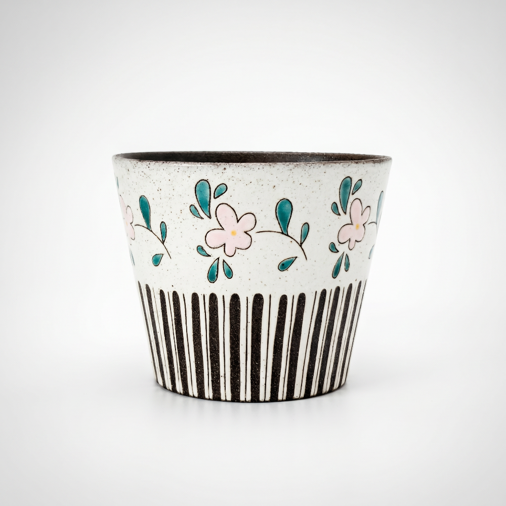 A straight-on studio shot of a tapered, handleless ceramic cup by Kove Ceramics on a clean white background. The speckled clay features a unique mixed pattern with hand-painted pink flowers and green leaves above bold, black vertical stripes, revealing a dark interior glaze.