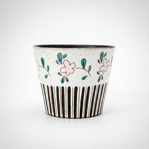 A straight-on studio shot of a tapered, handleless ceramic cup by Kove Ceramics on a clean white background. The speckled clay features a unique mixed pattern with hand-painted pink flowers and green leaves above bold, black vertical stripes, revealing a dark interior glaze.