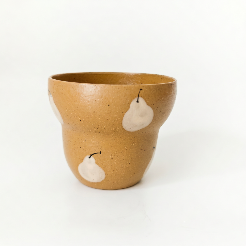 A slightly elevated studio shot of a stepped ceramic cup by Kove Ceramics, casting a soft shadow on a white surface. The image highlights the glossy mustard yellow speckled glaze and the charming hand-painted white pear motif.