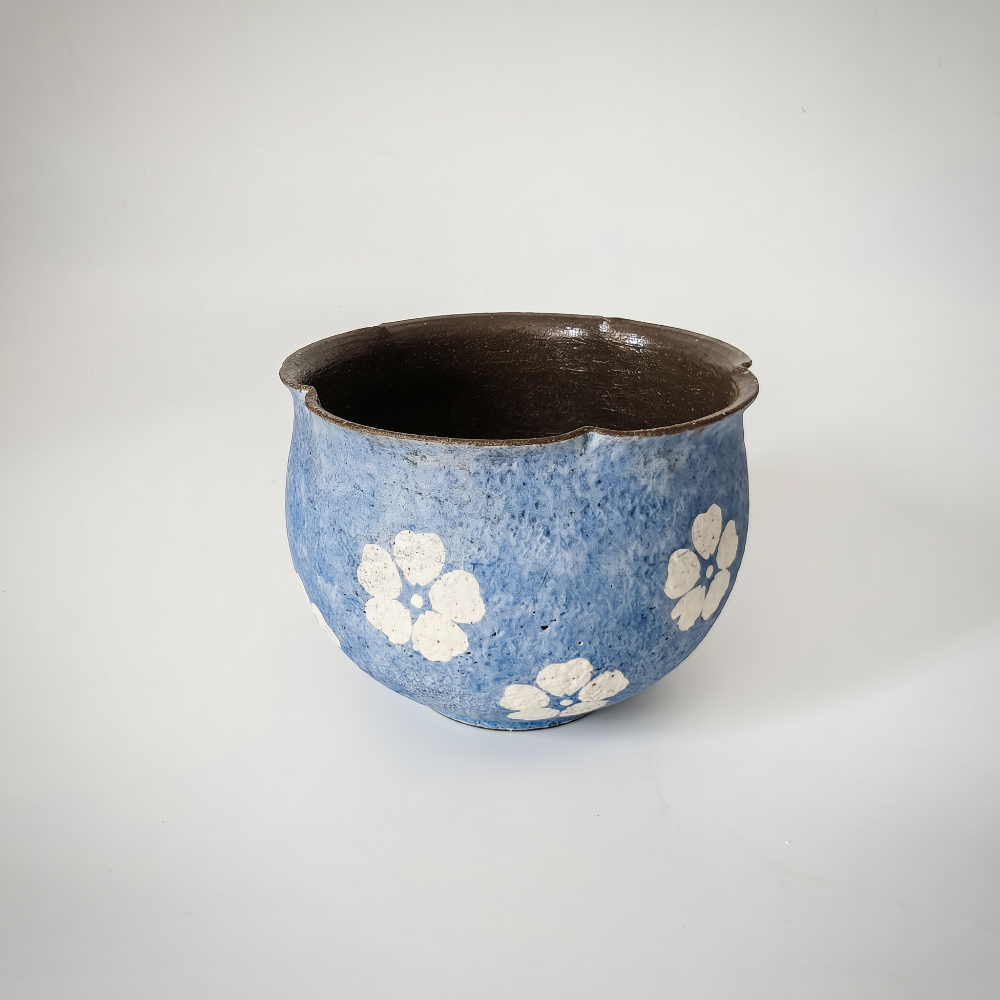 A high-angle studio shot of a handleless ceramic tea bowl by Kove Ceramics, specifically revealing its smooth, dark brown interior. The exterior features a textured, mottled blue glaze with white hand-painted blossoms, contrasted by a distinctively asymmetrical and wavy dark raw rim, set against a clean white background.