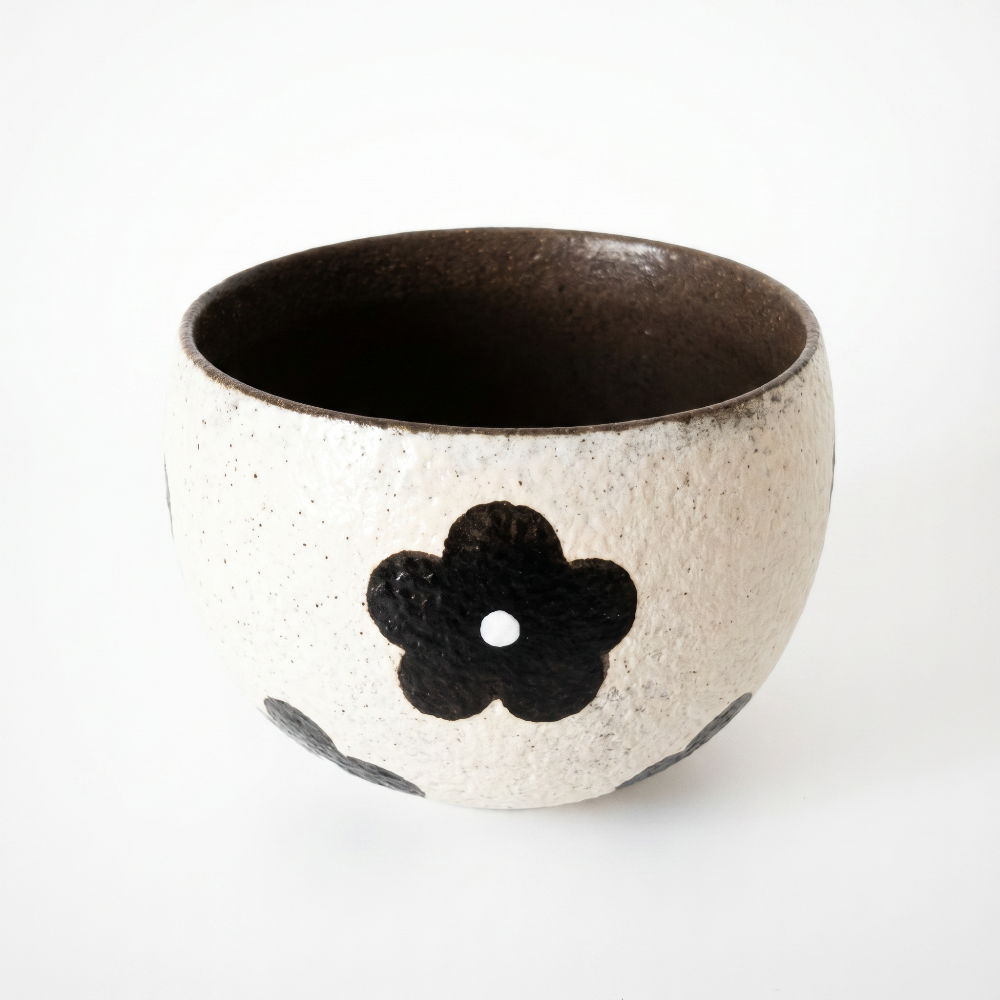 A high-angle view of a handmade speckled clay cup by Kove Ceramics, looking down into its deep, dark-glazed interior, contrasted by a textured exterior with a bold black retro floral motif