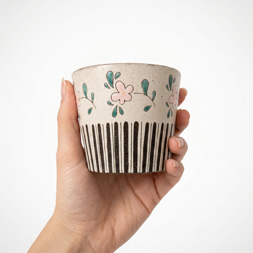 A hand holding a charming, handmade handleless ceramic cup by Kove Ceramics. The cup features a speckled cream clay body decorated with delicate hand-painted pink flowers and green leaves on the top half, contrasted by bold black vertical stripes on the bottom.