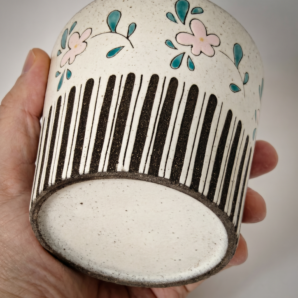 An in-hand shot showing the tilted bottom base of a handmade ceramic cup by Kove Ceramics. The view reveals the unglazed foot ring, the raw texture of the speckled clay, and the clean bottom edge of the black vertical stripes.
