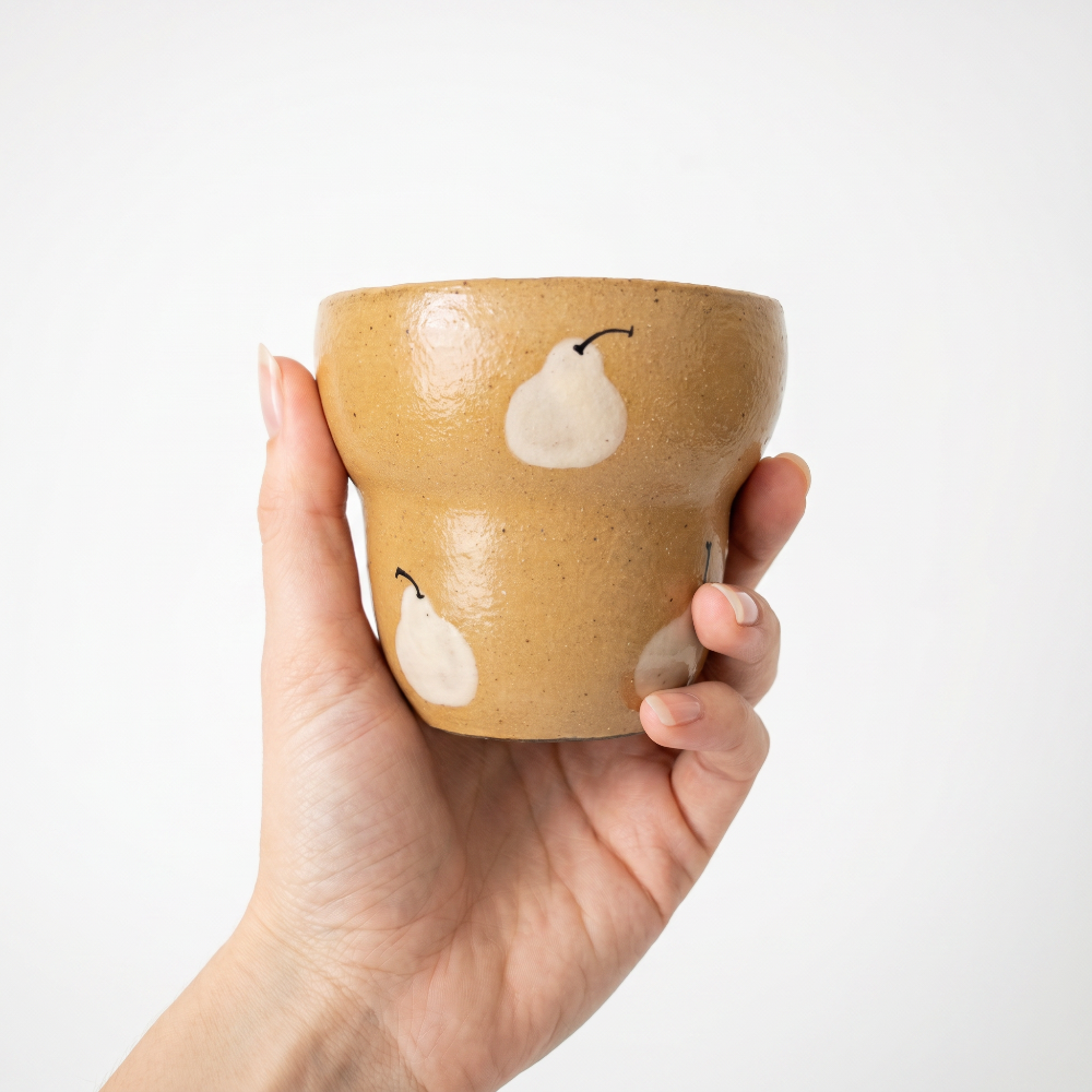 A hand holding a glossy mustard yellow ceramic cup by Kove Ceramics. The handleless cup features a unique stepped shape and is decorated with charming hand-painted white pears with black stems against a bright white background.