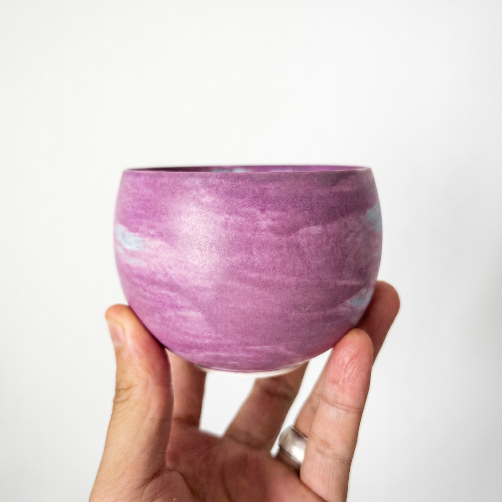 A close-up of a hand gently supporting a round, handleless ceramic tea bowl by Kove Ceramics, highlighting its smooth, matte finish and beautiful lavender and white watercolor swirling glaze.