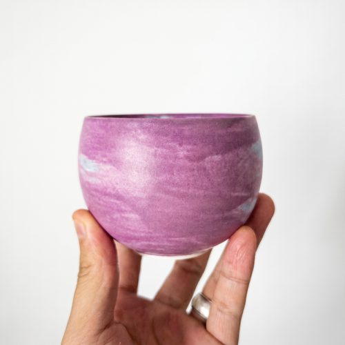 A close-up of a hand gently supporting a round, handleless ceramic tea bowl by Kove Ceramics, highlighting its smooth, matte finish and beautiful lavender and white watercolor swirling glaze.