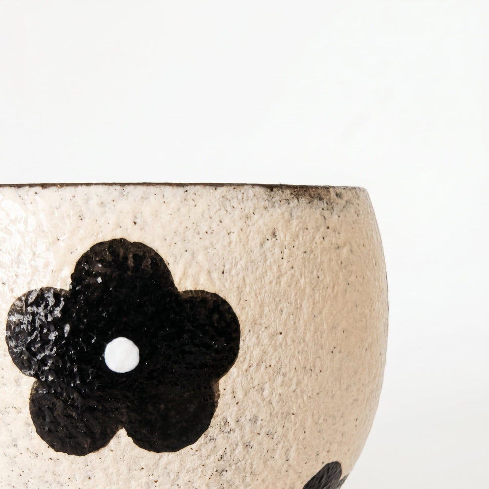 An extreme macro close-up of a ceramic cup by Kove Ceramics, highlighting the coarse, textured speckled cream surface and the authentic hand-painted details of a bold black retro flower with a white center.