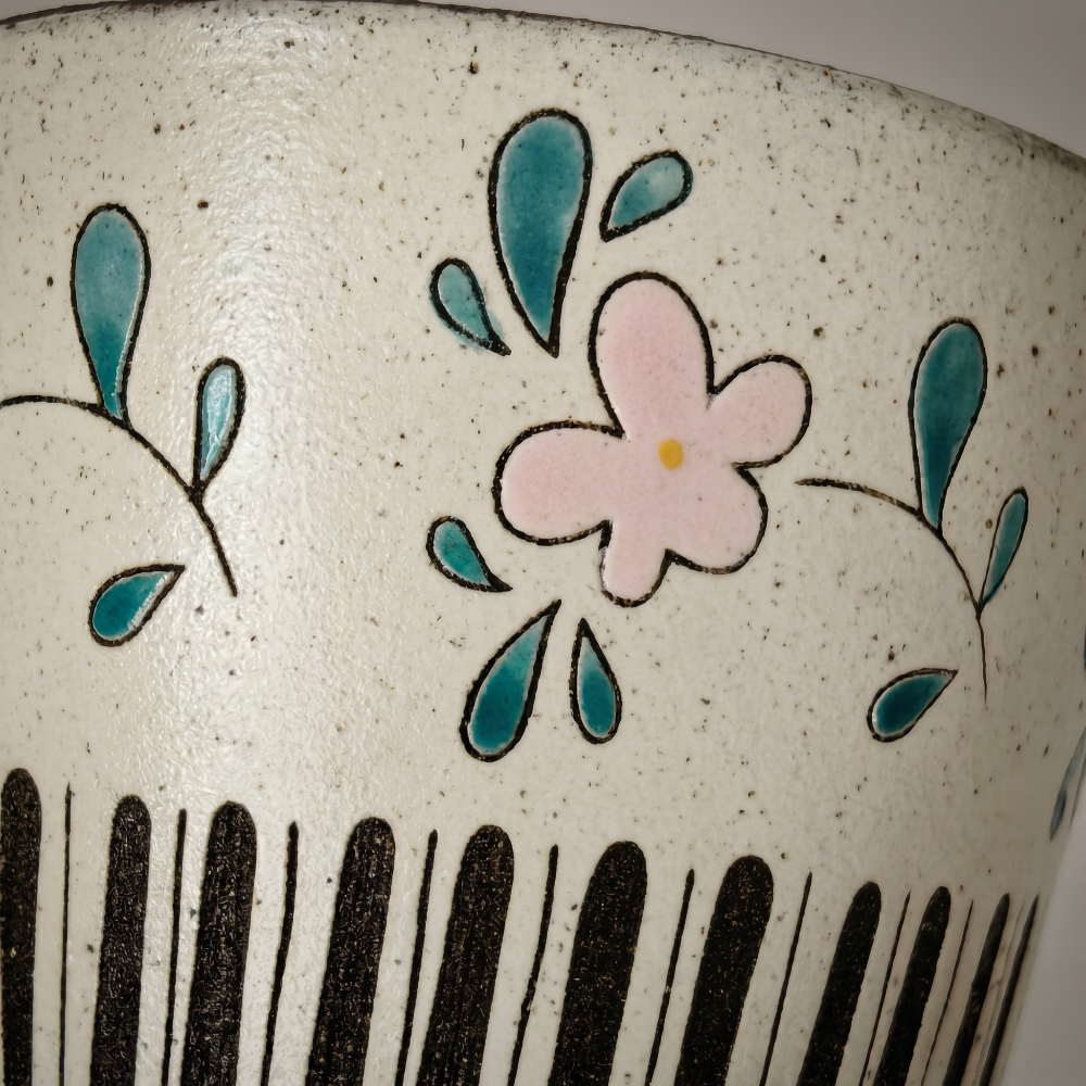 An extreme macro close-up of a handmade cup by Kove Ceramics, vividly revealing the raised texture of the hand-painted pink flower and teal leaves, the delicate brushstrokes of the black outlines, and the natural earthy speckles embedded in the clay body.