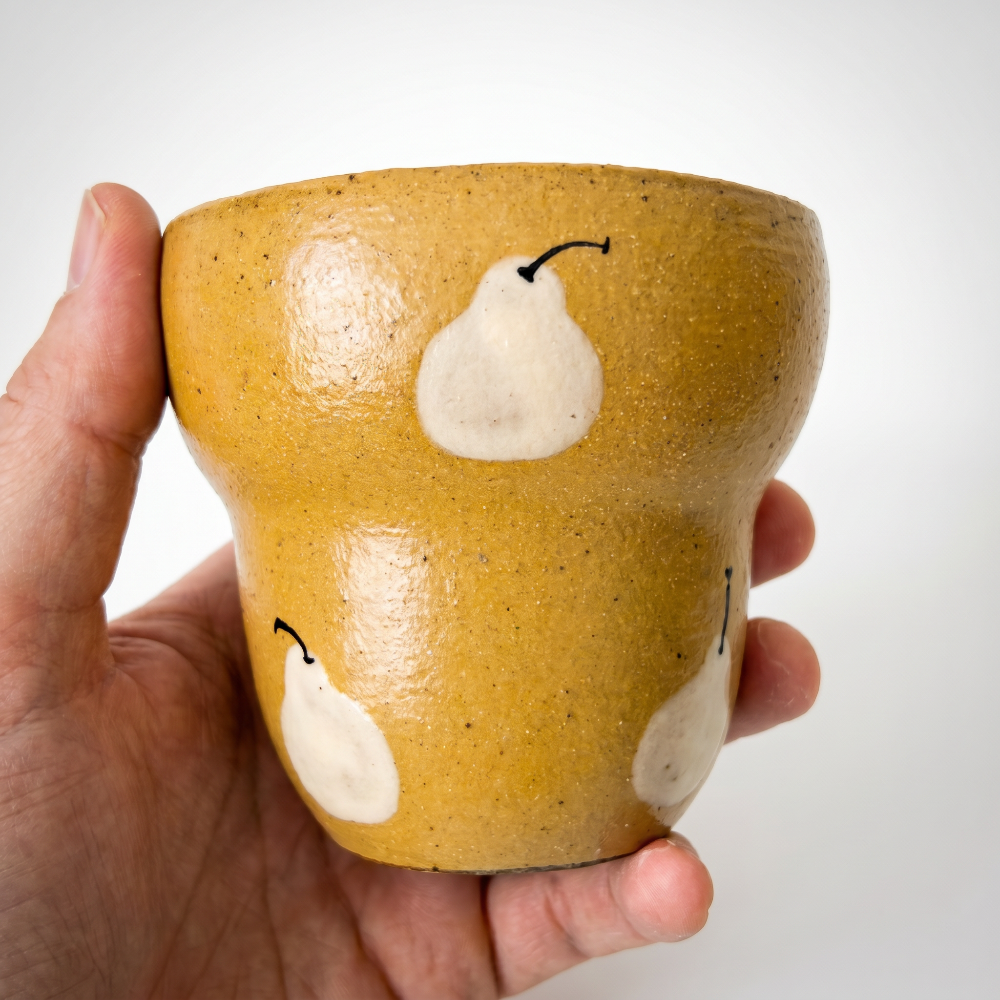 A tight macro close-up of a hand holding a stepped ceramic cup by Kove Ceramics. The detailed shot beautifully captures the high-gloss mustard yellow finish, the rustic clay speckles, and the slightly raised, tactile texture of the hand-painted white pear motif.