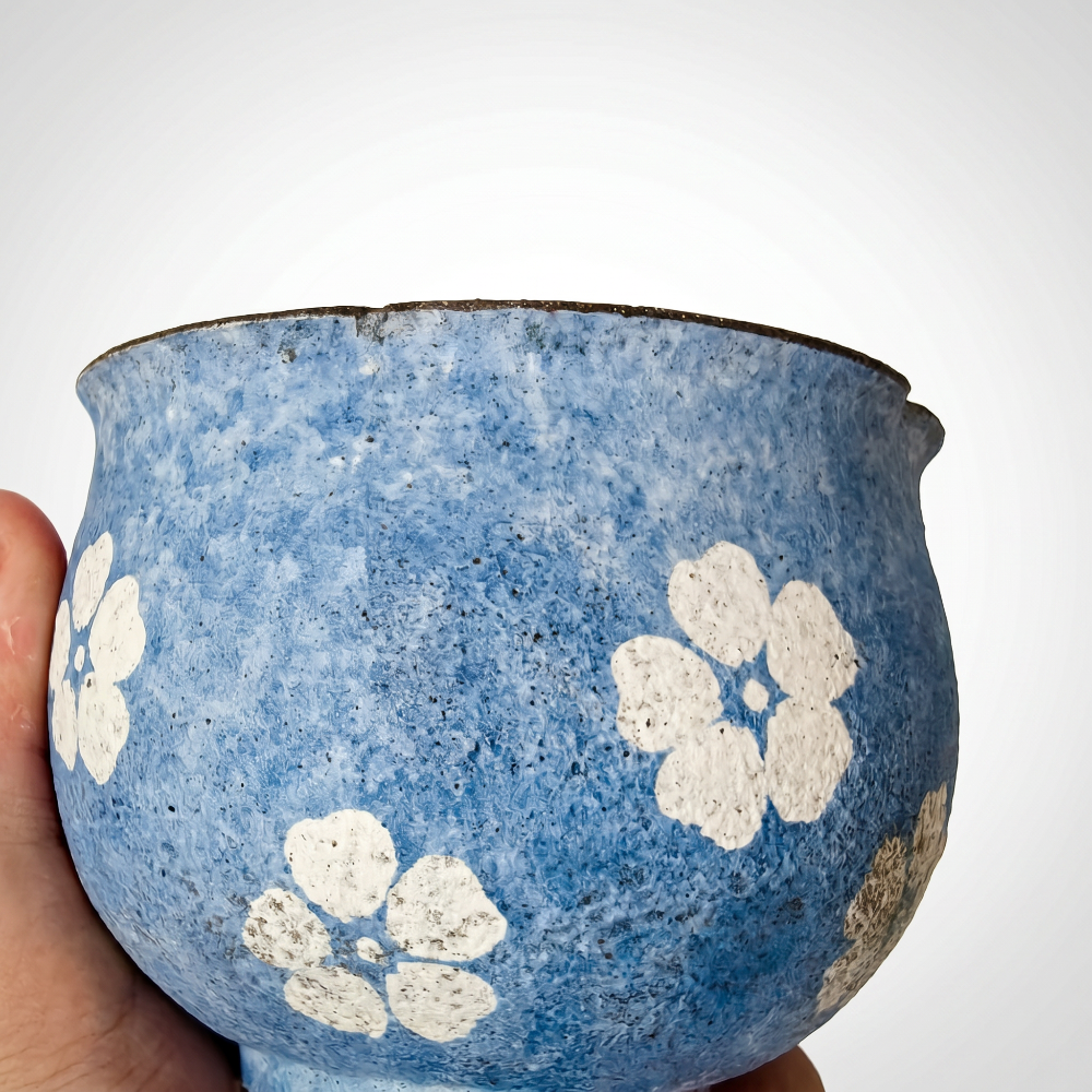 A handmade, round handleless ceramic An extreme macro close-up of a handmade blue ceramic cup by Kove Ceramics, partially held by a hand. The tight shot highlights the rough, stone-like mottled blue surface and the thick, raised tactile details of the hand-painted white floral motifs, finished with a dark raw rim.