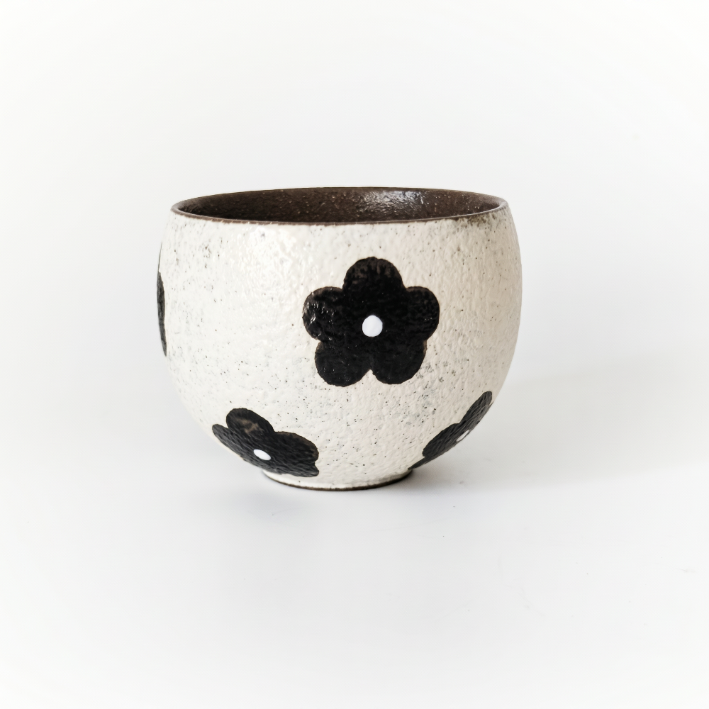 A minimalist studio shot of an artisan handmade ceramic handleless cup by Kove Ceramics, showcasing a speckled white body with hand-painted black retro flowers and a dark interior, resting on a clean white surface.