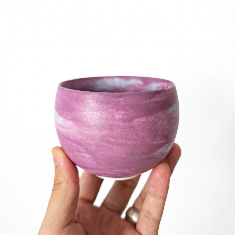 A hand holding a round, handmade ceramic handleless matcha cup by Kove Ceramics, showcasing a vibrant purple and white marbled watercolor glaze against a bright white background