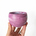 A hand holding a round, handmade ceramic handleless matcha cup by Kove Ceramics, showcasing a vibrant purple and white marbled watercolor glaze against a bright white background