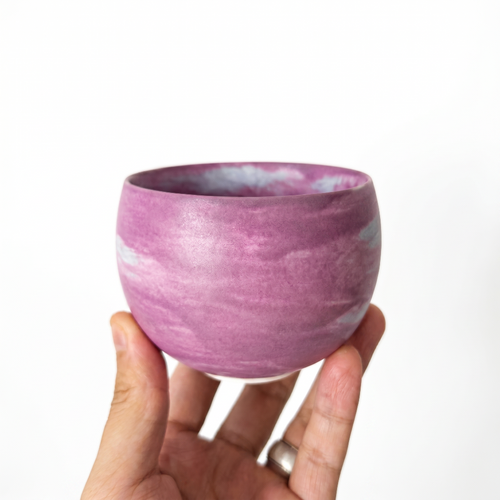 A hand holding a round, handmade ceramic handleless matcha cup by Kove Ceramics, showcasing a vibrant purple and white marbled watercolor glaze against a bright white background