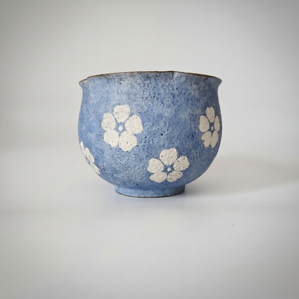 A handmade, round handleless ceramic cup by Kove Ceramics resting on a white surface with soft shadows. It features a rustic, highly textured blue stone-like glaze, decorated with hand-painted white blossoms, and is finished with a dark, subtly wavy raw rim.