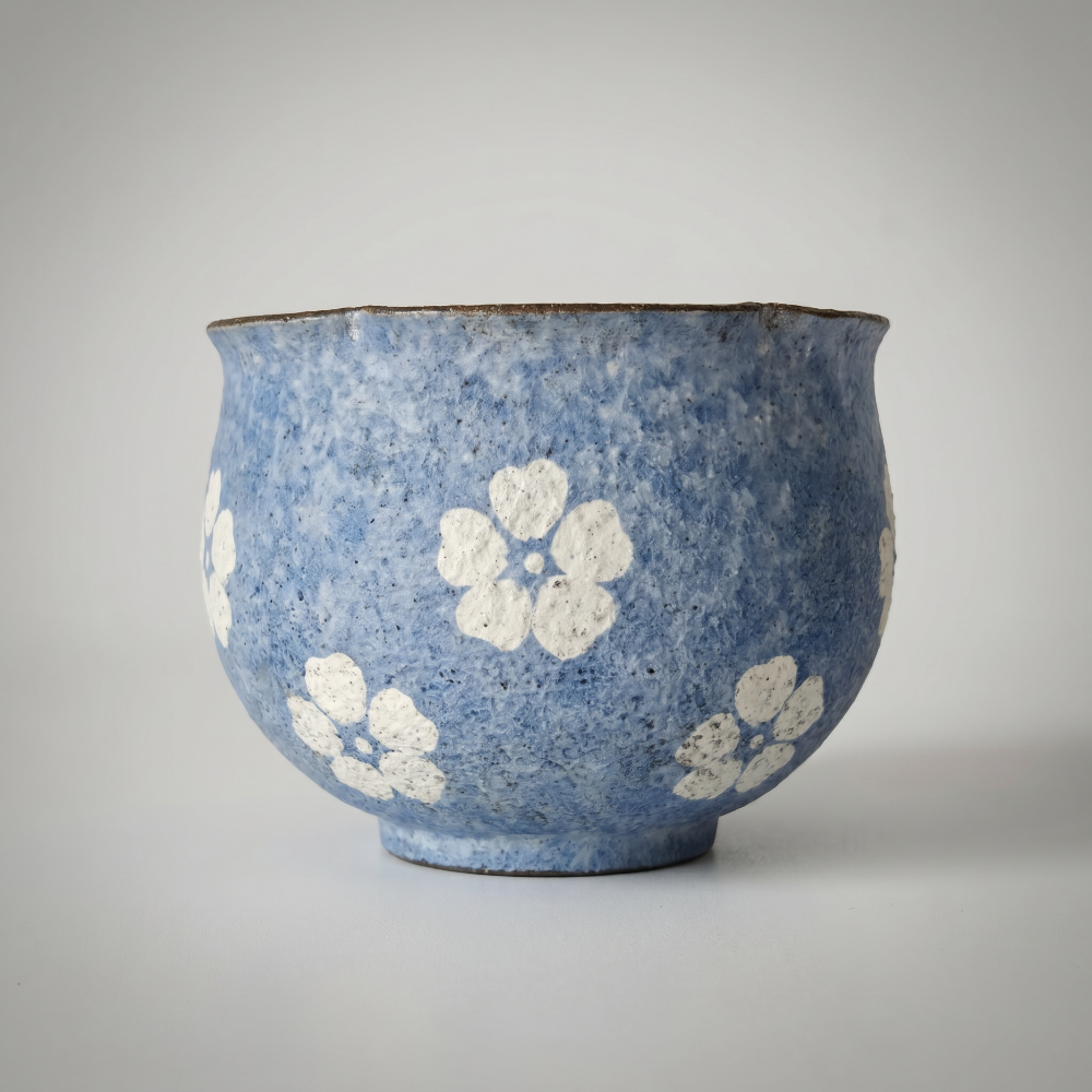 A soft-lit still-life photo of a round, handleless ceramic cup by Kove Ceramics. The cup features a textured, mottled blue glaze with hand-painted white blossoms and a dark uneven rim, casting a gentle shadow to the right against a muted grey-white background.