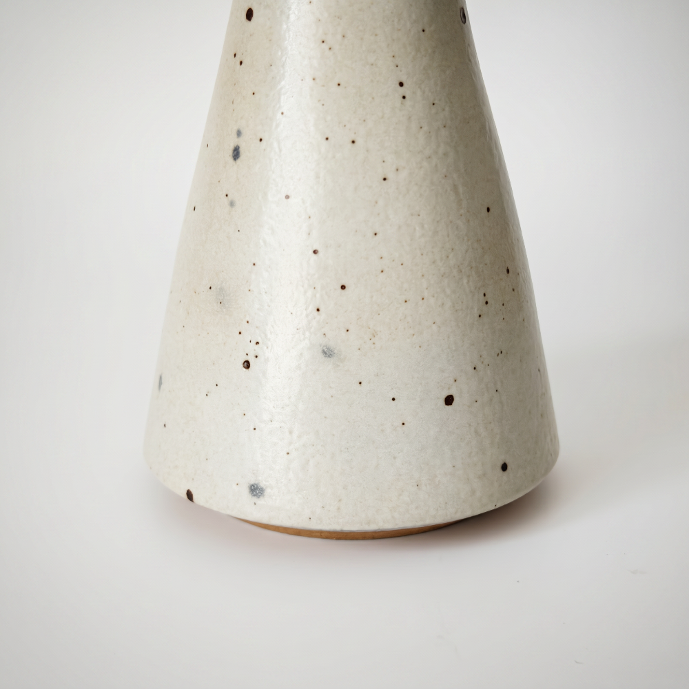 Close-up detail of the conical vase base by Kove Ceramics, highlighting the tactile cream glaze with organic dark speckles and a glimpse of the natural unglazed clay foot.