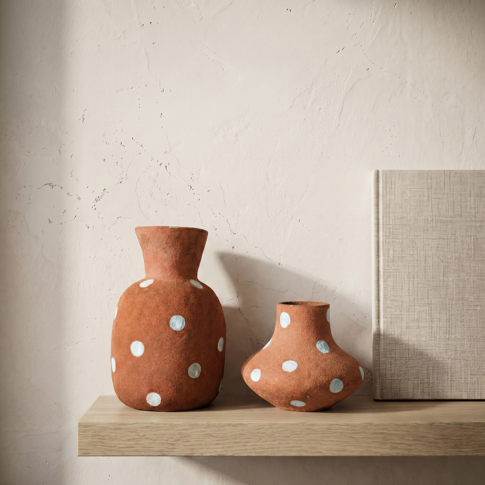 A lifestyle styling shot of two rustic terracotta polka dot vases by Kove Ceramics, beautifully displayed on a light wooden floating shelf against a textured plaster wall in warm sunlight.