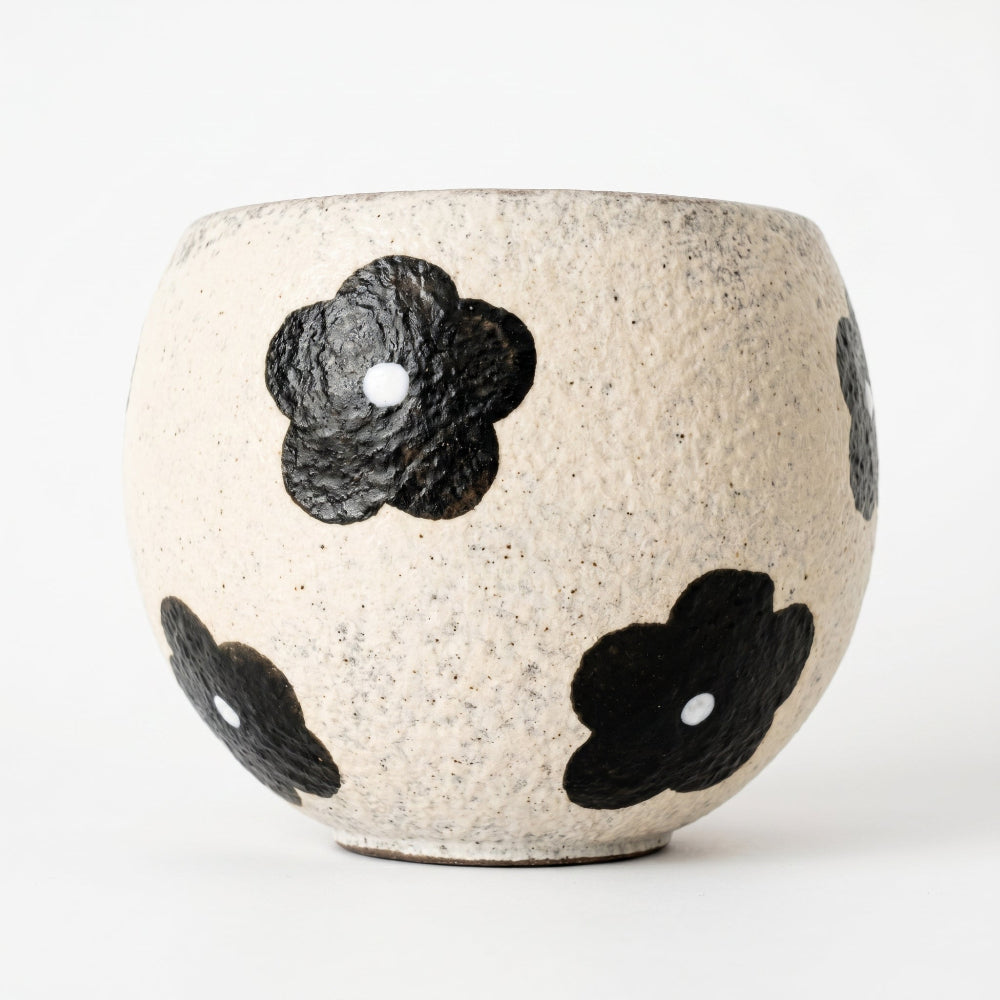 A round, handleless ceramic matcha bowl by Kove Ceramics, showcasing a highly textured speckled cream clay surface decorated with bold, rustic black floral patterns against a clean white background.