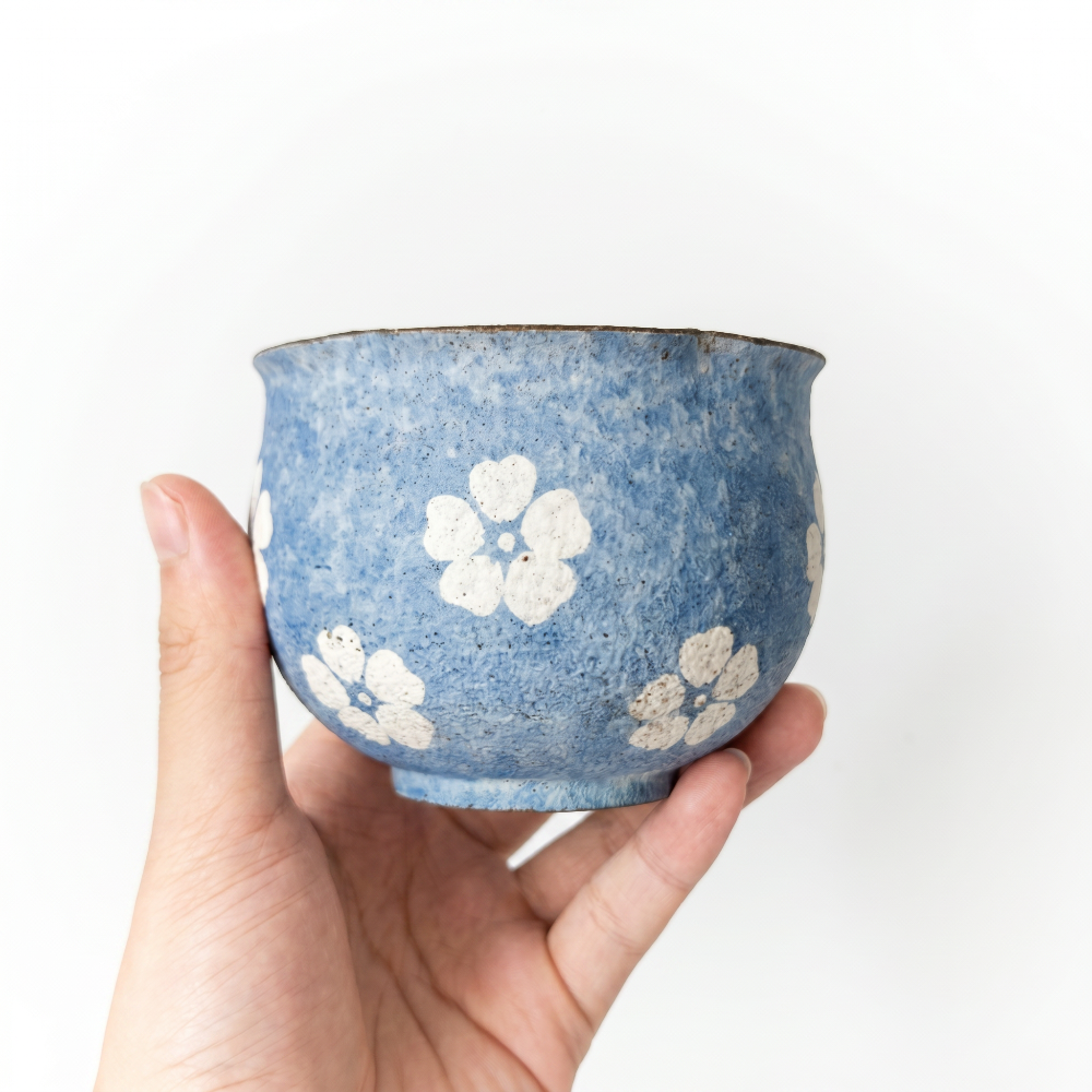 A close-up of a hand holding a handleless ceramic cup by Kove Ceramics. The cup features a highly textured, mottled blue glaze with a slightly flared rim, decorated with charming hand-painted white floral motifs against a clean white background.