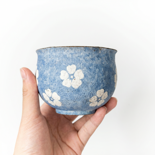 A close-up of a hand holding a handleless ceramic cup by Kove Ceramics. The cup features a highly textured, mottled blue glaze with a slightly flared rim, decorated with charming hand-painted white floral motifs against a clean white background.