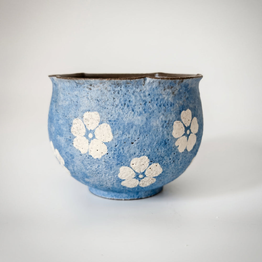 A straight-on studio shot of a handleless ceramic tea bowl by Kove Ceramics on a white background. The piece features a heavily textured, mottled blue glaze, hand-painted white flower motifs, a dark asymmetrical wavy rim, and a small footed base