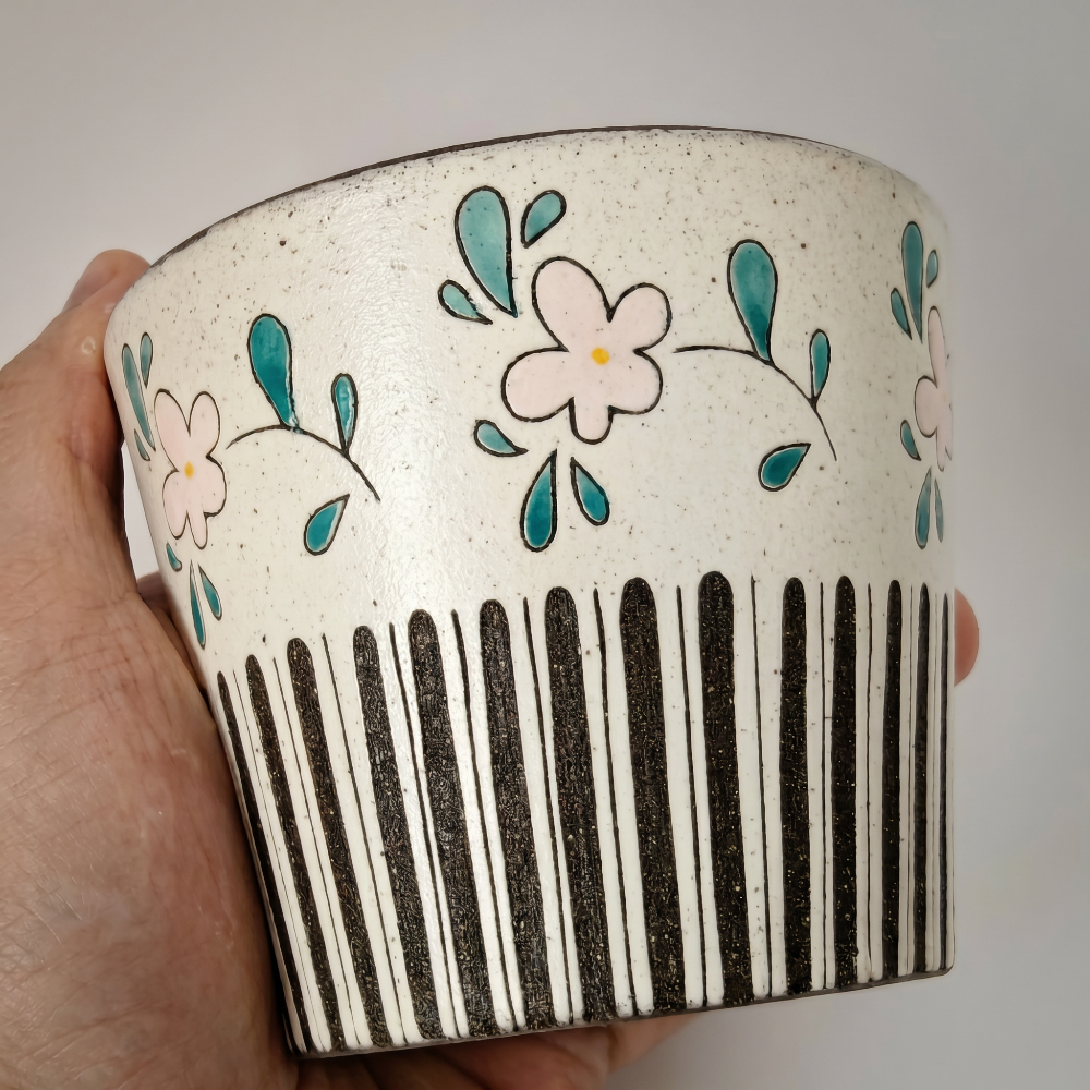 A tight close-up shot of a hand firmly holding a tapered ceramic cup by Kove Ceramics, highlighting the hand-feel, the subtle satin sheen on the speckled clay, and its whimsical hand-painted retro pink flowers and black stripes.