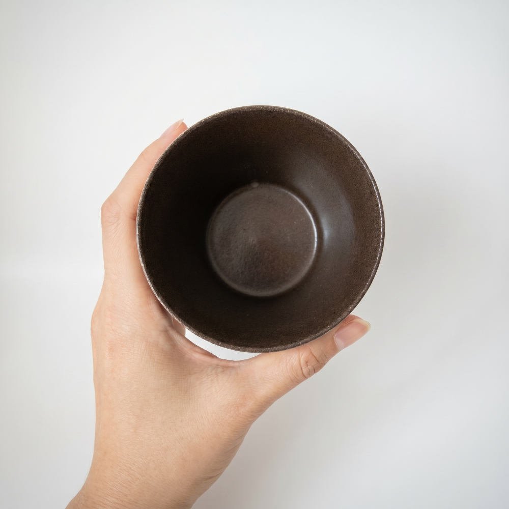 A top-down bird's-eye view of a hand holding a handmade ceramic cup by Kove Ceramics, revealing its perfectly round, thin rim and a deep, smooth dark brown glazed interior against a white background.