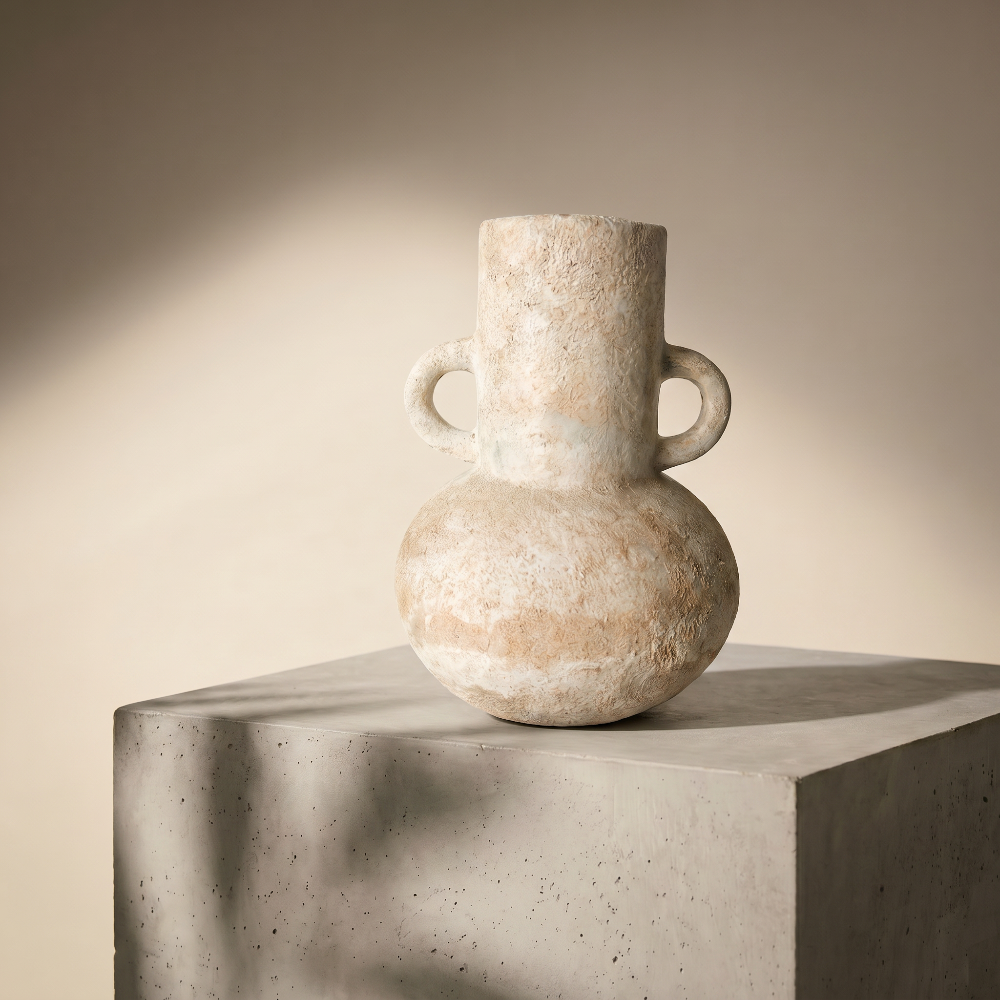 A sculptural, wabi-sabi style rustic ceramic vase by Kove Ceramics, featuring an earthy, distressed finish and double ring handles, elegantly displayed on a modern concrete pedestal bathed in warm, dappled sunlight.