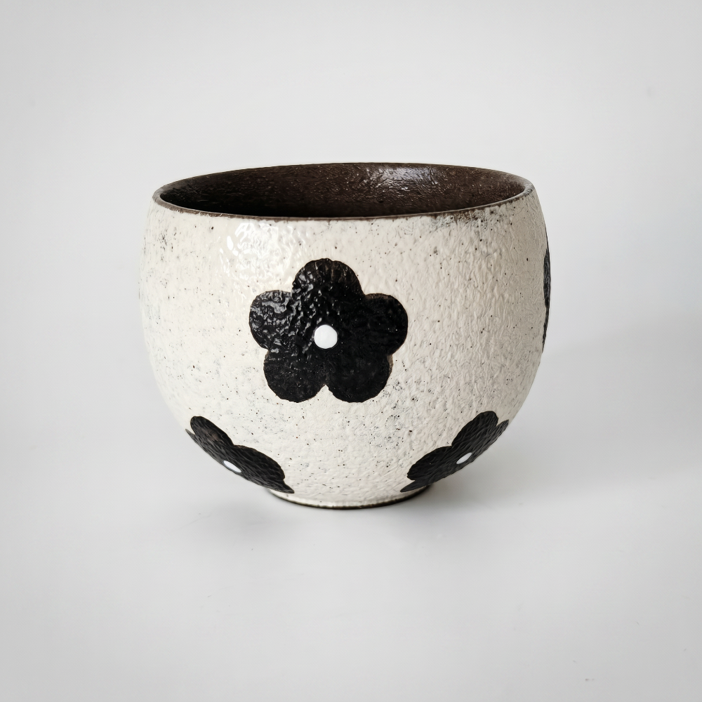 A rustic wabi-sabi stoneware tea tumbler by Kove Ceramics, resting on a white surface with soft shadows, showcasing its textured cream glaze and bold black hand-painted floral motifs.