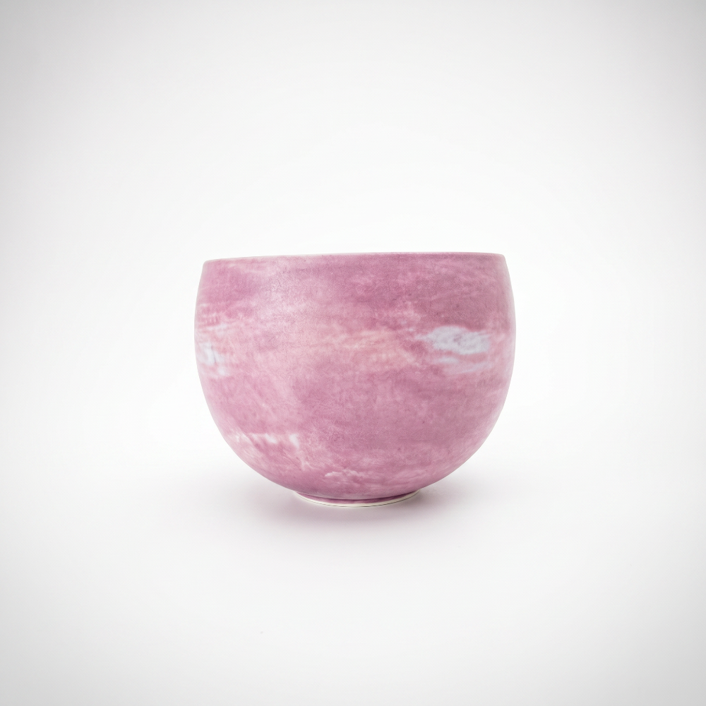 A clean, minimalist studio shot of a round, handleless pink ceramic matcha bowl by Kove Ceramics, resting on a white surface and showcasing its cloudy, marbled purple and white watercolor glaze.